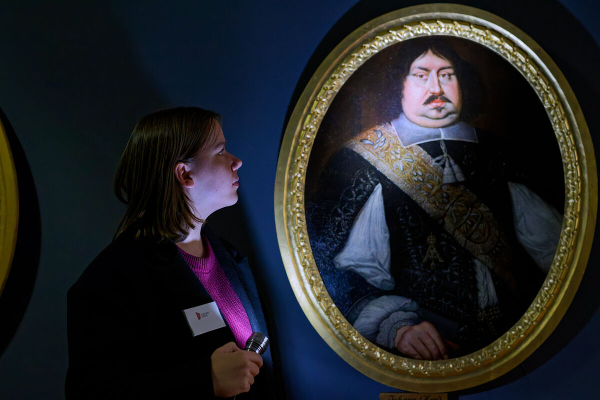 Eine Frau steht im Museum vor einer dunklen Wand mit Gemälden. Mit einer Taschenlampe beleuchtet sie ein Porträt im Schloss.